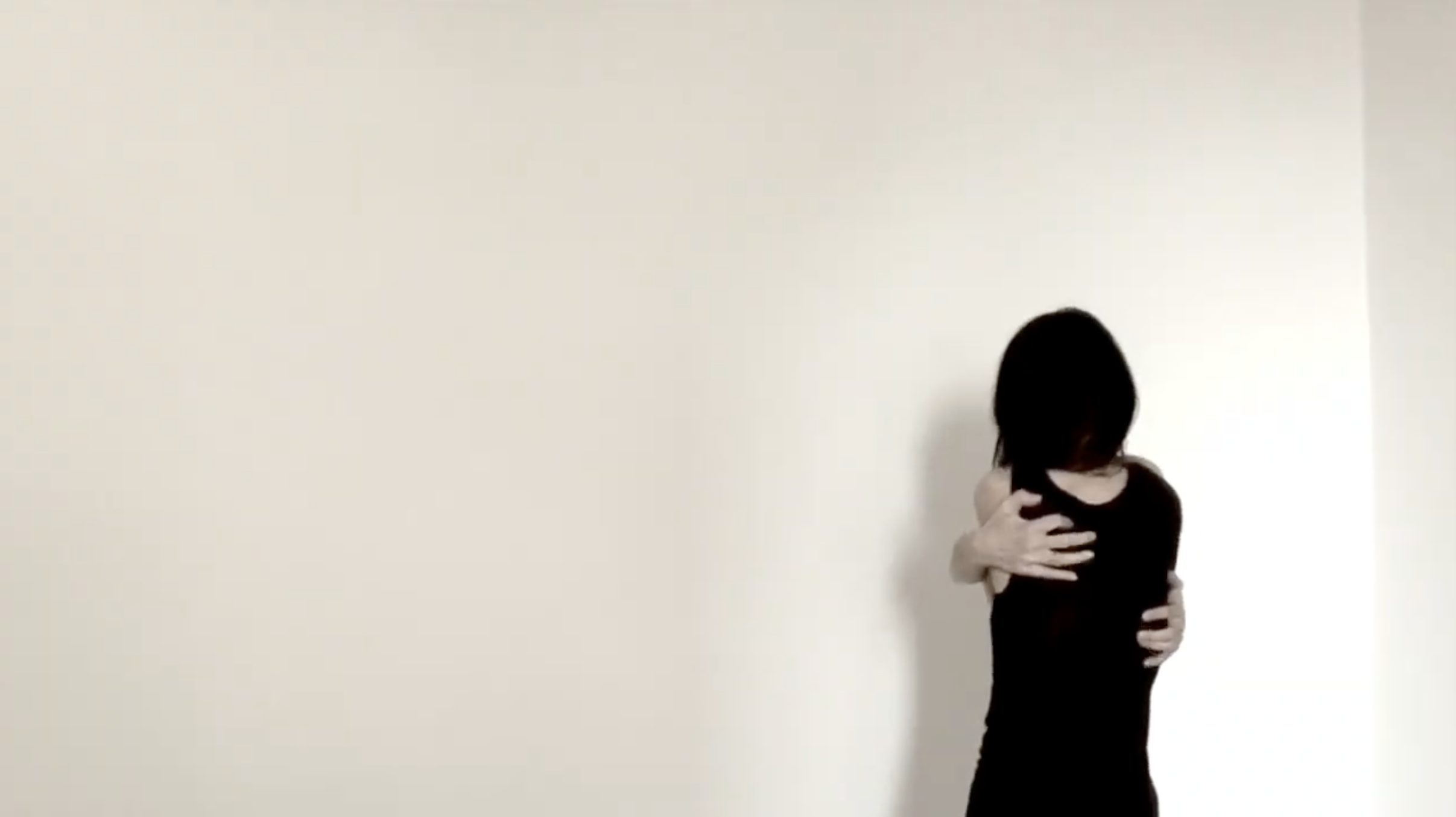 A woman with long black hair turned away from the camera, dressed in black, hugging herself.